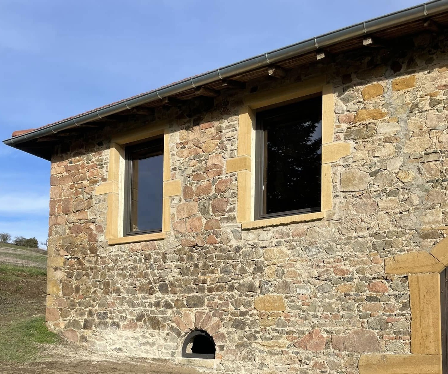 Maison en pierres dorées rénovée en Beaujolais — menuiseries sombres neuves, ciel bleu — réalisation suivie par Julien Parriaux AMO